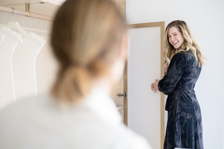 Two women in a light-filled room, one with blonde hair in a white top, the other with wavy blonde hair in a dark patterned robe, smiling and looking at each other.