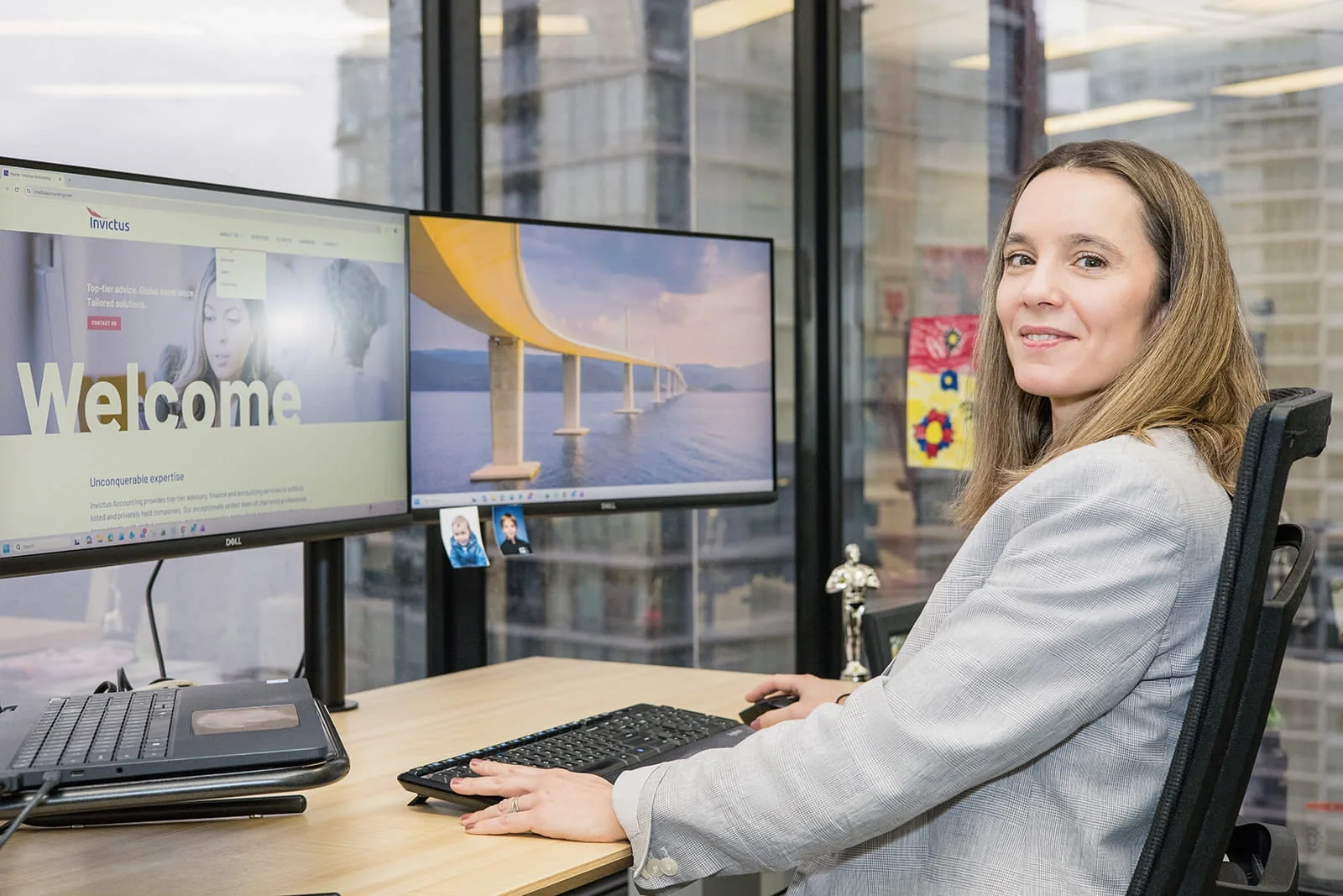 A woman in a light gray blazer working at a desk with two monitors, a keyboard, and a laptop, in an office with large windows showing a cityscape outside.