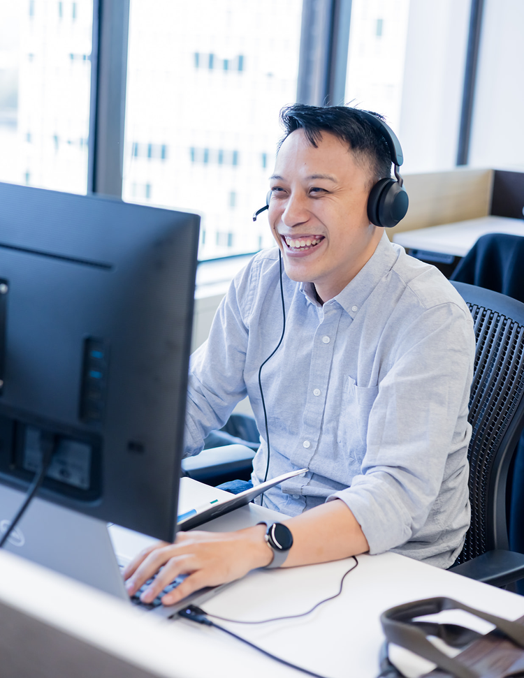 A young man wearing a headset and smiling while working at a computer in an office with large windows.