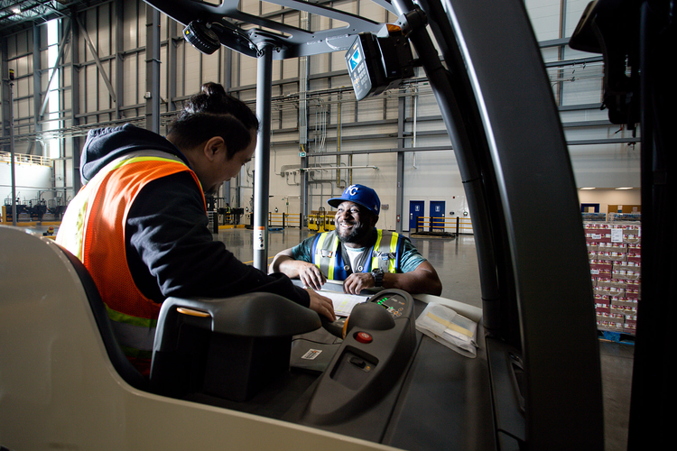 Two workers, one wearing a safety vest and sitting inside a forklift, the other smiling and wearing a baseball cap and safety vest, having a discussion inside a warehouse.