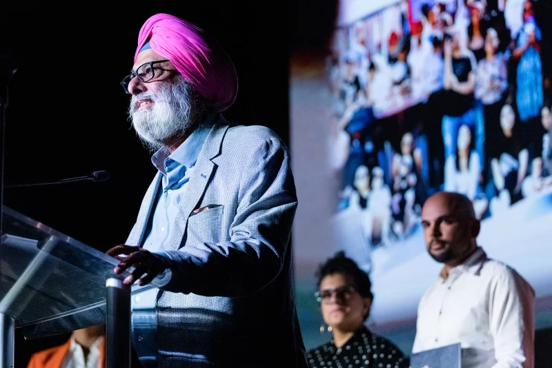 A man with a white beard and glasses wearing a pink turban and a checkered blazer is speaking at a podium during a presentation. Behind him, two individuals, one with glasses and a woman, are looking on. A large photo collage is visible in the background.