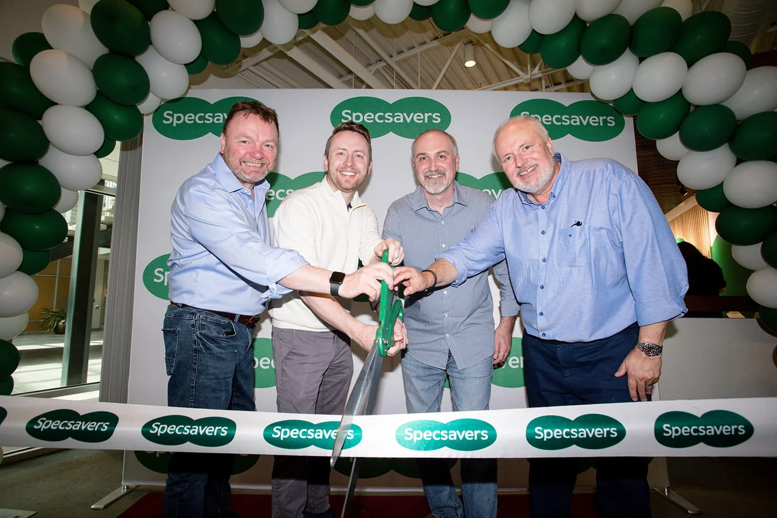 Five men at a ribbon-cutting event for Specsavers, with green and white balloons and a Specsavers backdrop. They are smiling and holding large green scissors.