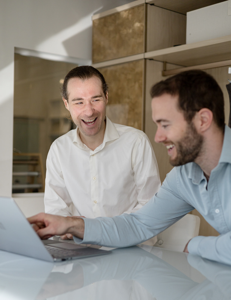 Two men are sitting at a table with a laptop, smiling and laughing during a discussion or meeting.