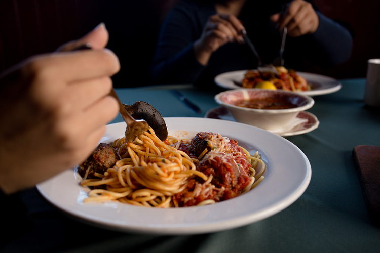 Person eating spaghetti with meatballs at a dining table, with other plates of food and a bowl of sauce visible in the background.