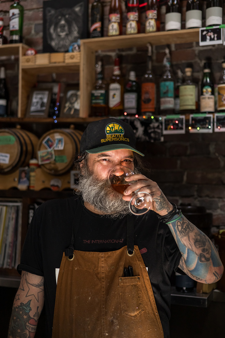 A man with a beard and tattoos enjoying a drink in a bar, wearing a Seattle SuperSonics cap and a brown apron, with bottles on shelves behind him.