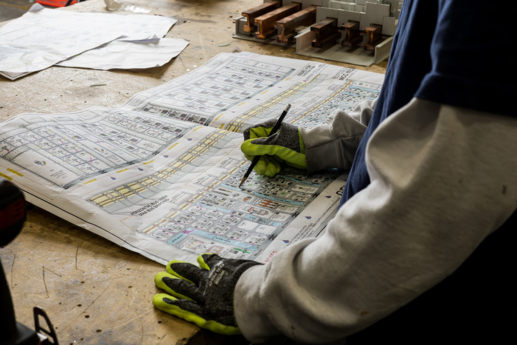 A person wearing gloves and a jacket is examining a detailed construction or architectural blueprint on a workbench, with scattered papers and small wooden structures nearby.