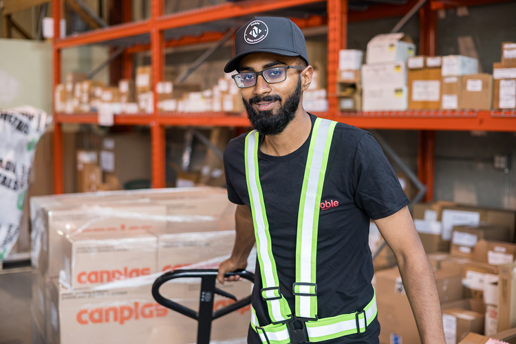 A man with glasses and a black cap working in a warehouse, wearing a high-visibility safety vest, standing in front of shelves filled with boxes.