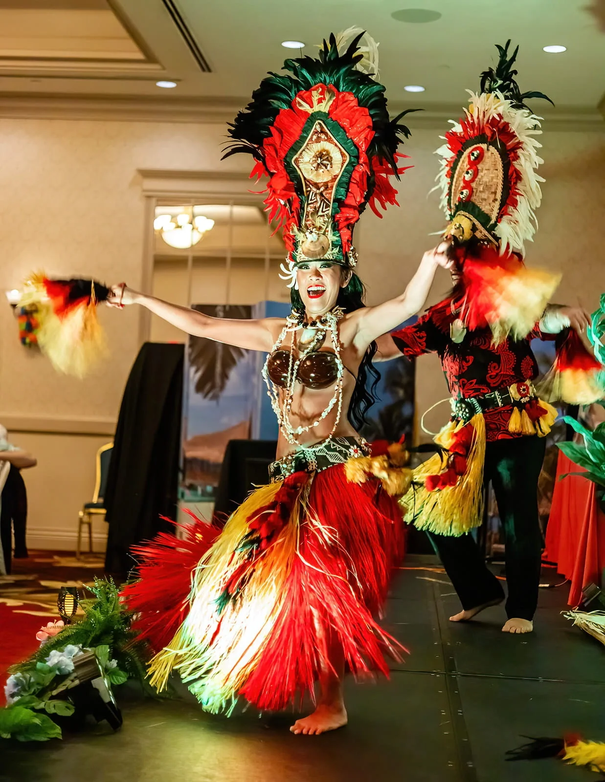 A woman performing a traditional Polynesian dance, wearing a colorful feathered headdress and skirt, with a decorated top, on stage.