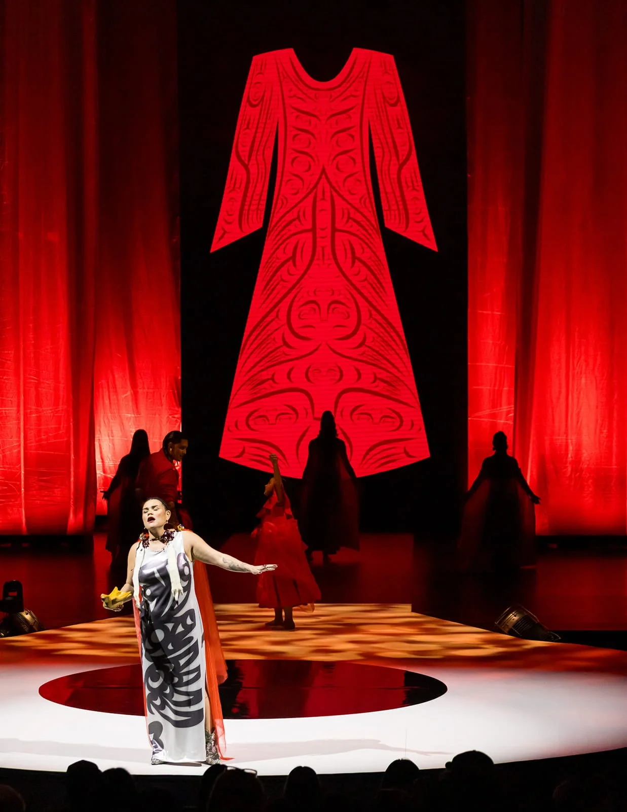A woman performing on stage in front of a red and black backdrop with a large indigenous design. Several dancers in traditional attire are in the background, with a red curtain on either side of the stage.