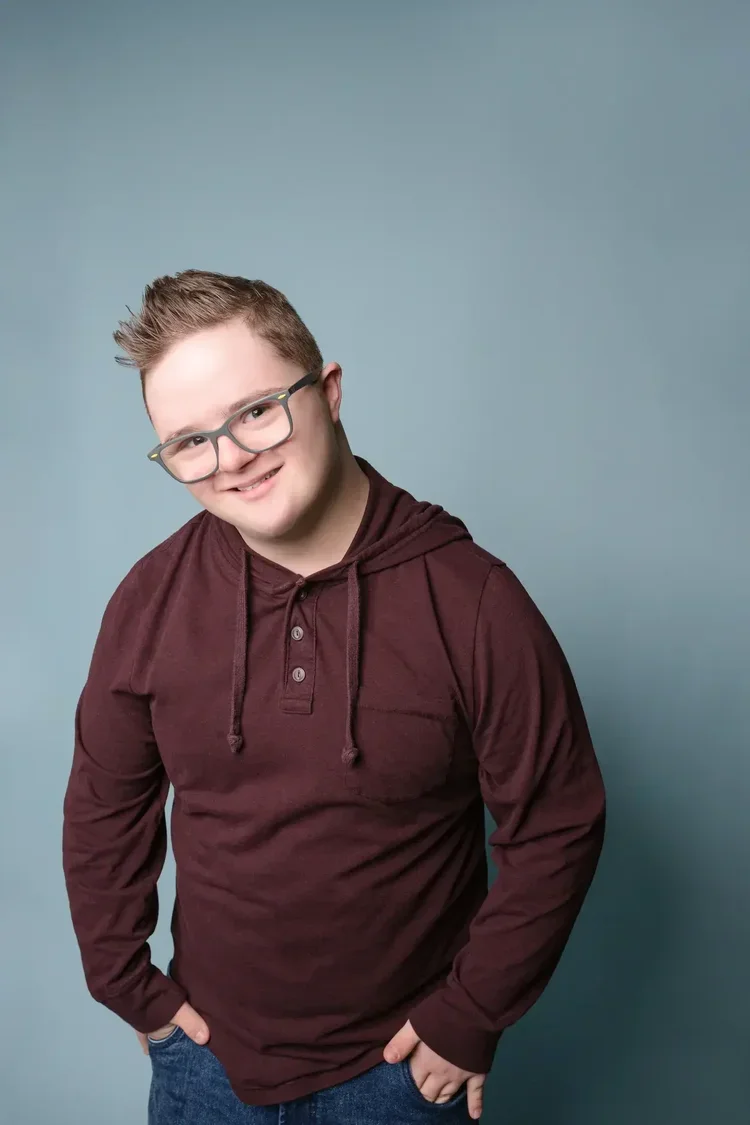 A smiling young man with glasses, wearing a maroon hoodie and jeans, standing with one hand in his pocket against a plain blue background.