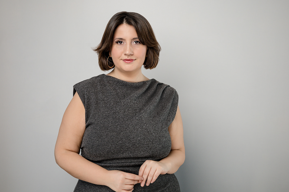 A woman with shoulder-length brown hair wearing a dark gray sleeveless dress stands against a light gray background.