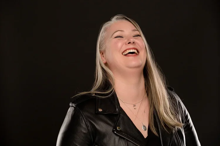 A woman with long blonde hair wearing a black leather jacket, laughing with her eyes closed against a dark background.