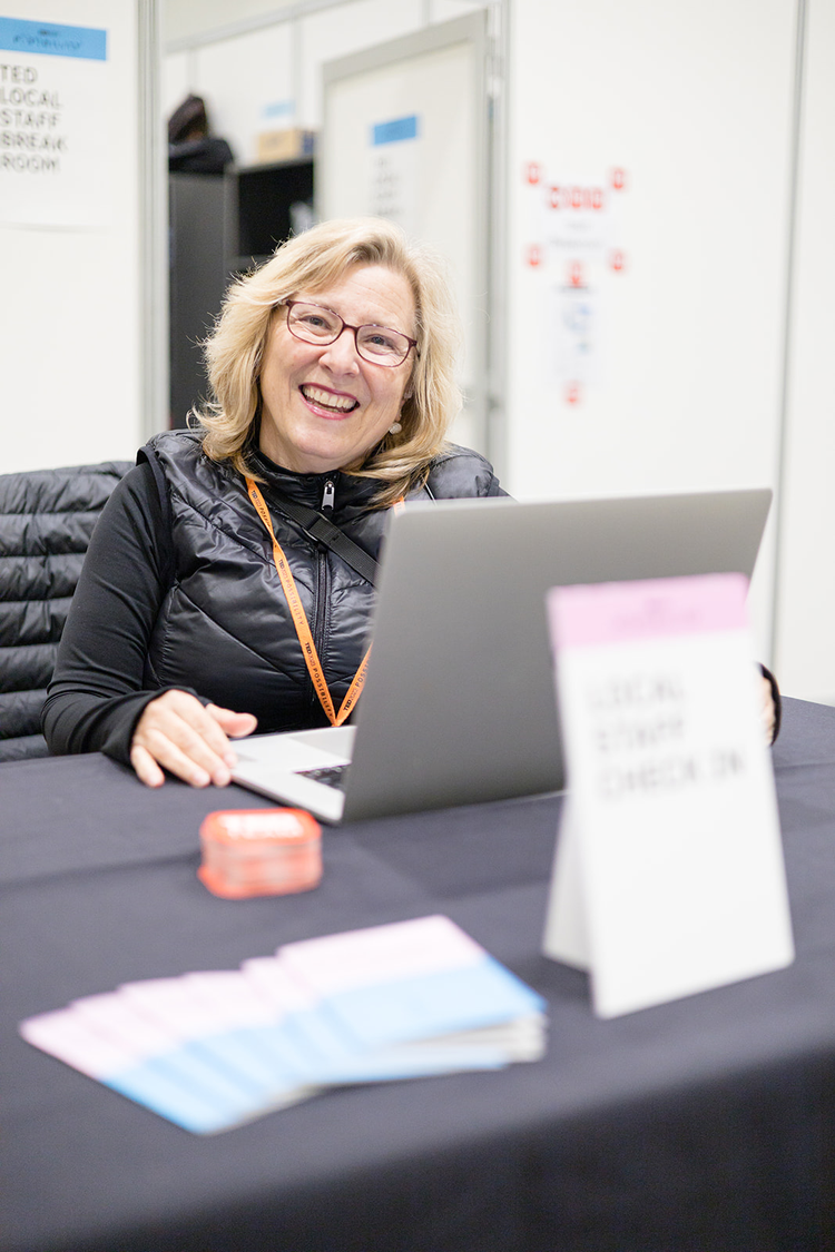 Smiling woman with glasses sitting at a table with a laptop in an indoor setting, possibly at a conference or event, with pamphlets and a name tag on the table.