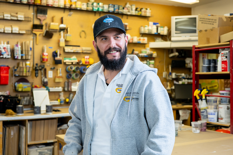 A man with a beard wearing a gray hoodie and a black baseball cap standing inside a hardware store or workshop with tools and supplies on the shelves in the background.