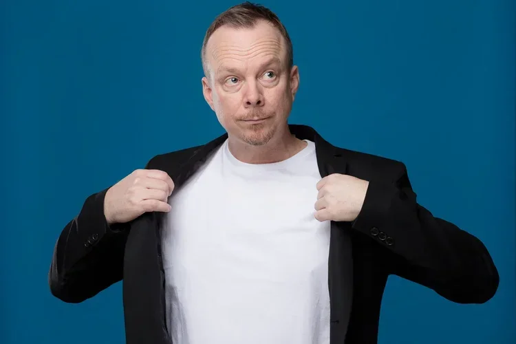 A man with short red hair and a goatee opening a black blazer to reveal a plain white shirt against a plain blue background.