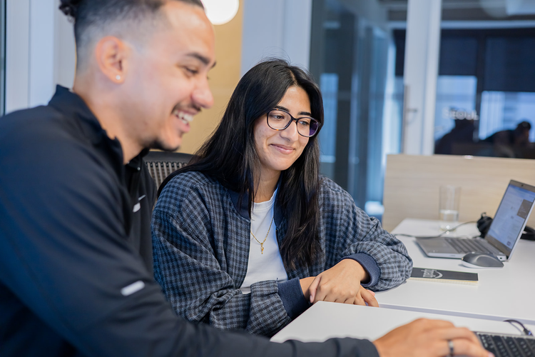 Two young adults sitting at a table in an office, smiling and looking at a laptop screen.