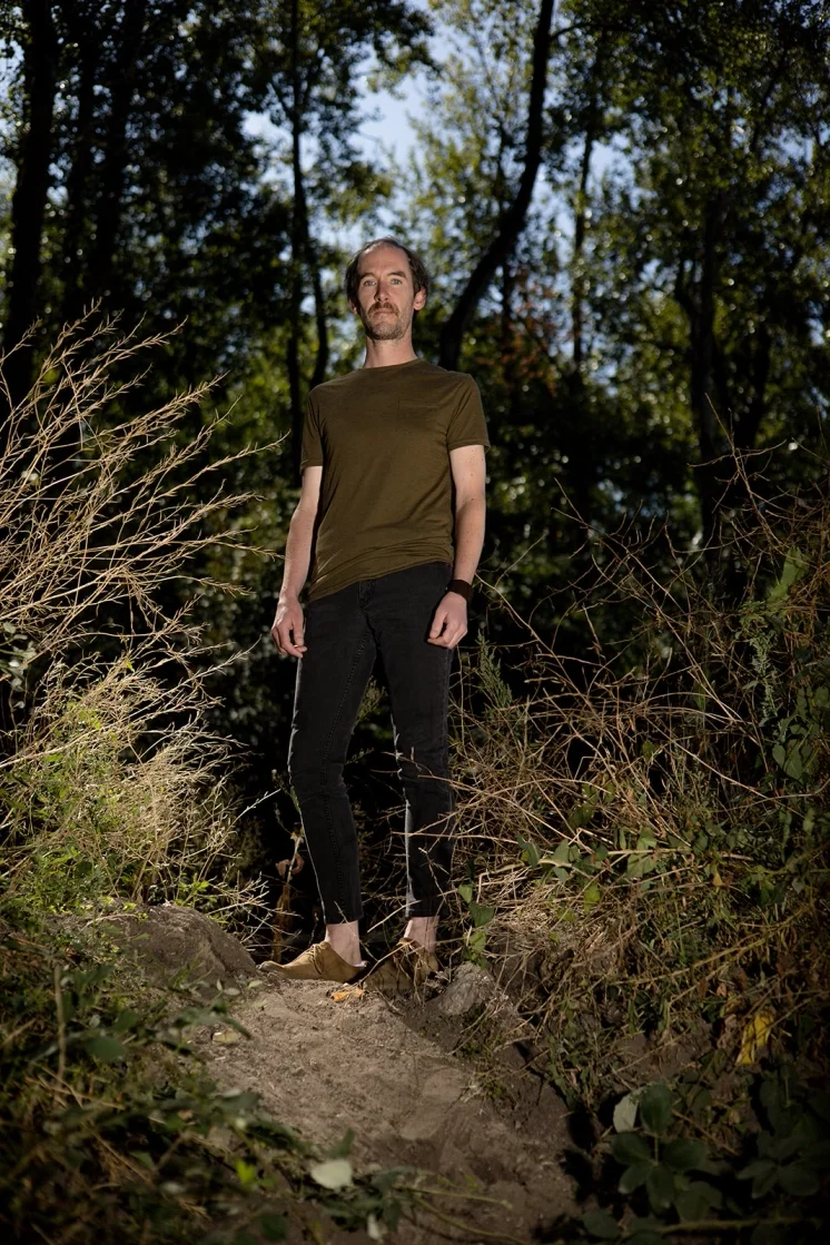 Man standing in a wooded outdoor area with trees and vegetation around him, wearing a green t-shirt, black pants, and tan shoes.