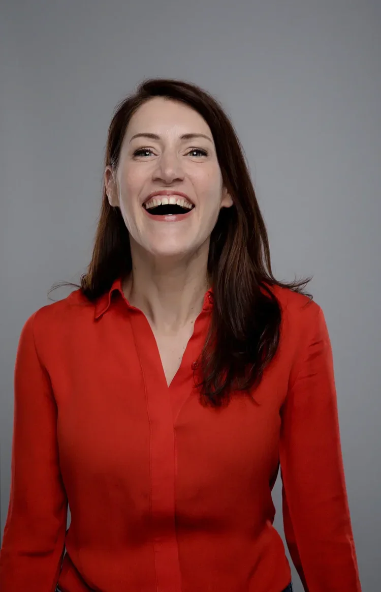 A woman with test skin and long dark brown hair, smiling and wearing a red blouse, standing against a solid gray background.