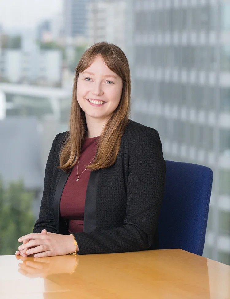 A woman with long light brown hair, wearing a black blazer and maroon top, smiling and seated at a wooden table in a modern office with large windows and city buildings in the background.