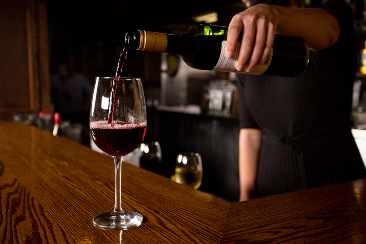 A person pouring red wine into a glass at a bar counter.