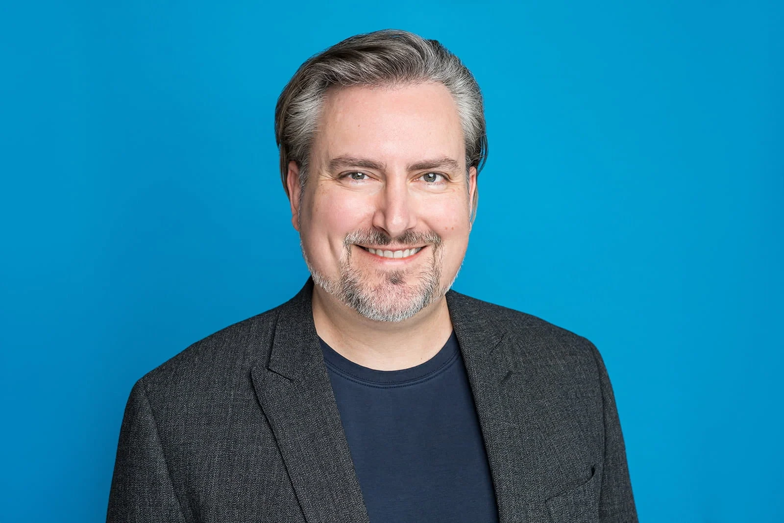 Portrait of a middle-aged man with gray hair and beard, smiling, wearing a dark blazer and navy shirt, against a blue background.