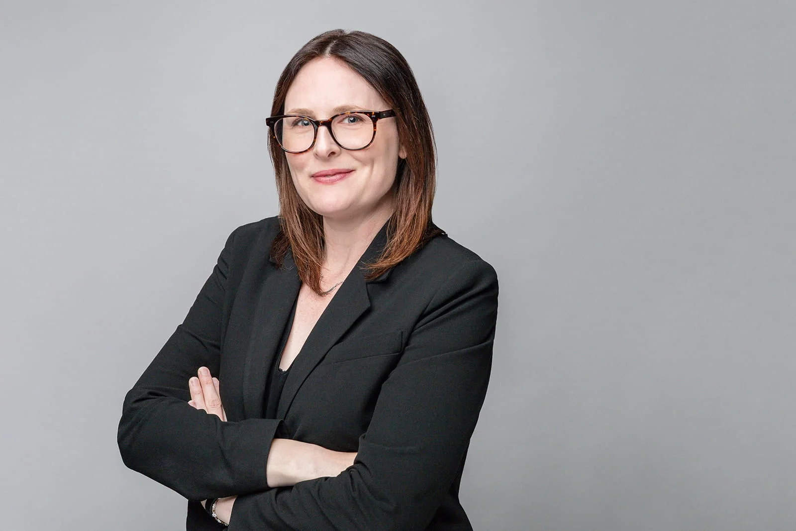 A professional woman with shoulder-length brown hair and glasses, wearing a black blazer, standing with arms crossed against a plain gray background.