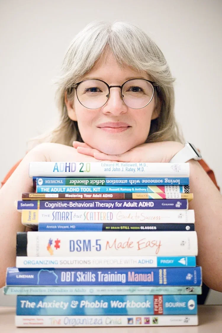A woman with gray hair and glasses is resting her chin on her hands, which are placed on a stack of books about ADHD and mental health.