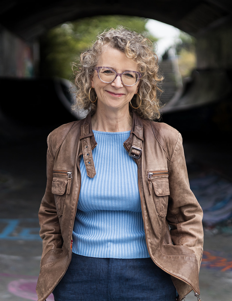 Middle-aged woman with curly blonde hair, wearing glasses, gold hoop earrings, a blue ribbed sweater, and a brown leather jacket, standing outdoors near a tunnel with graffiti on the walls.