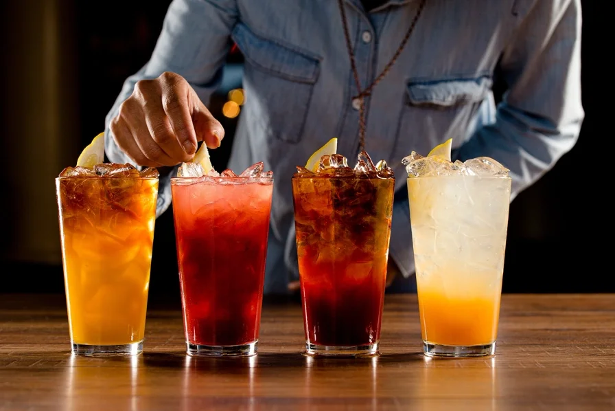A person wearing a denim shirt adding lemon slices to colorful iced drinks on a wooden table.