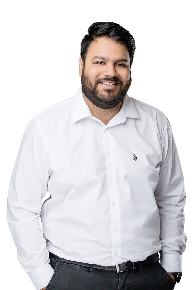 A smiling man with dark hair and a beard wearing a white button-down shirt and black pants against a white background.