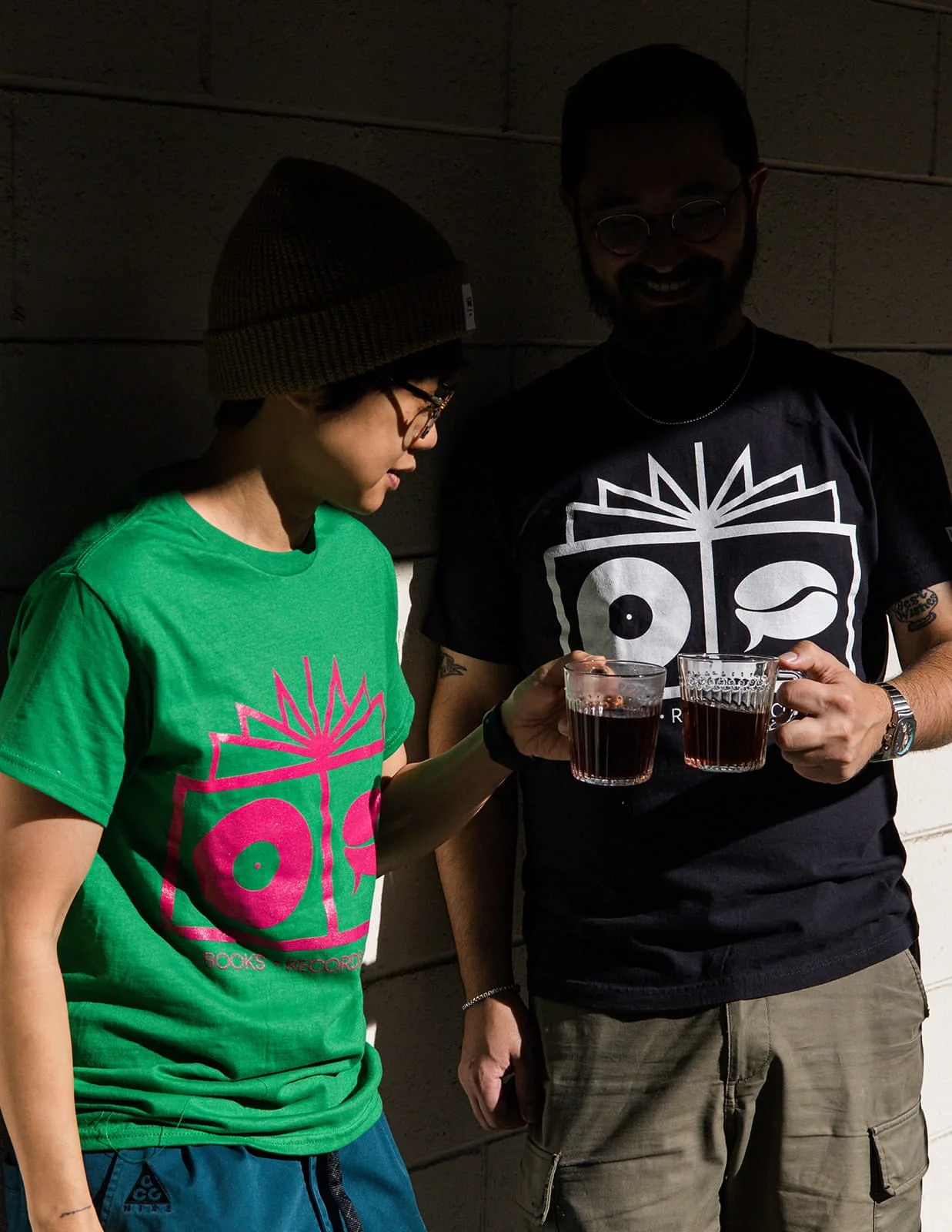 Two people wearing matching black and green T-shirts with a stylized face design, holding glasses of dark red beverage and smiling.