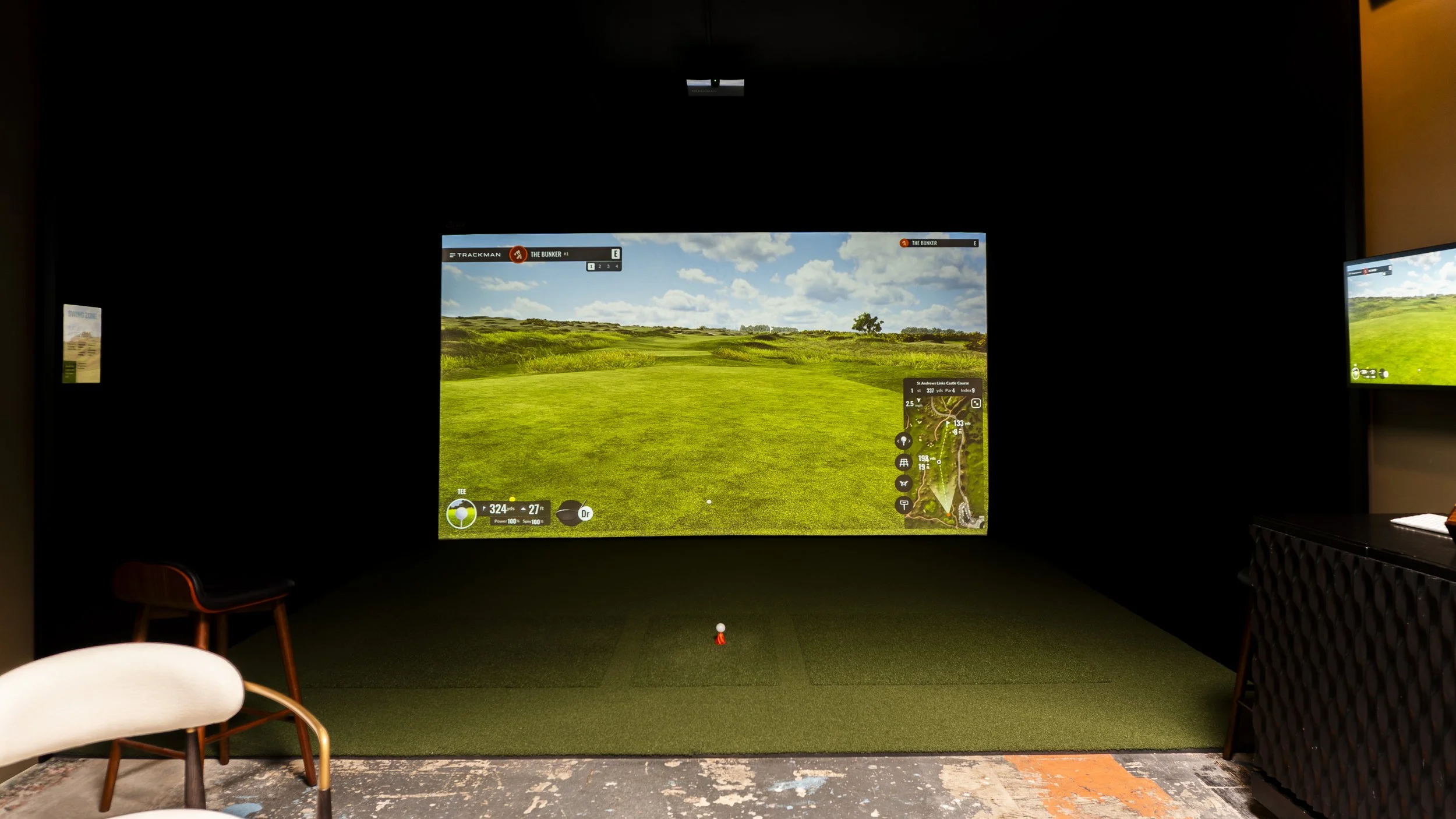 Indoor golf simulator displaying a virtual golf course with green grass, trees, and blue sky with clouds. Surrounding dark walls, chairs in the foreground, and screens on the sides.