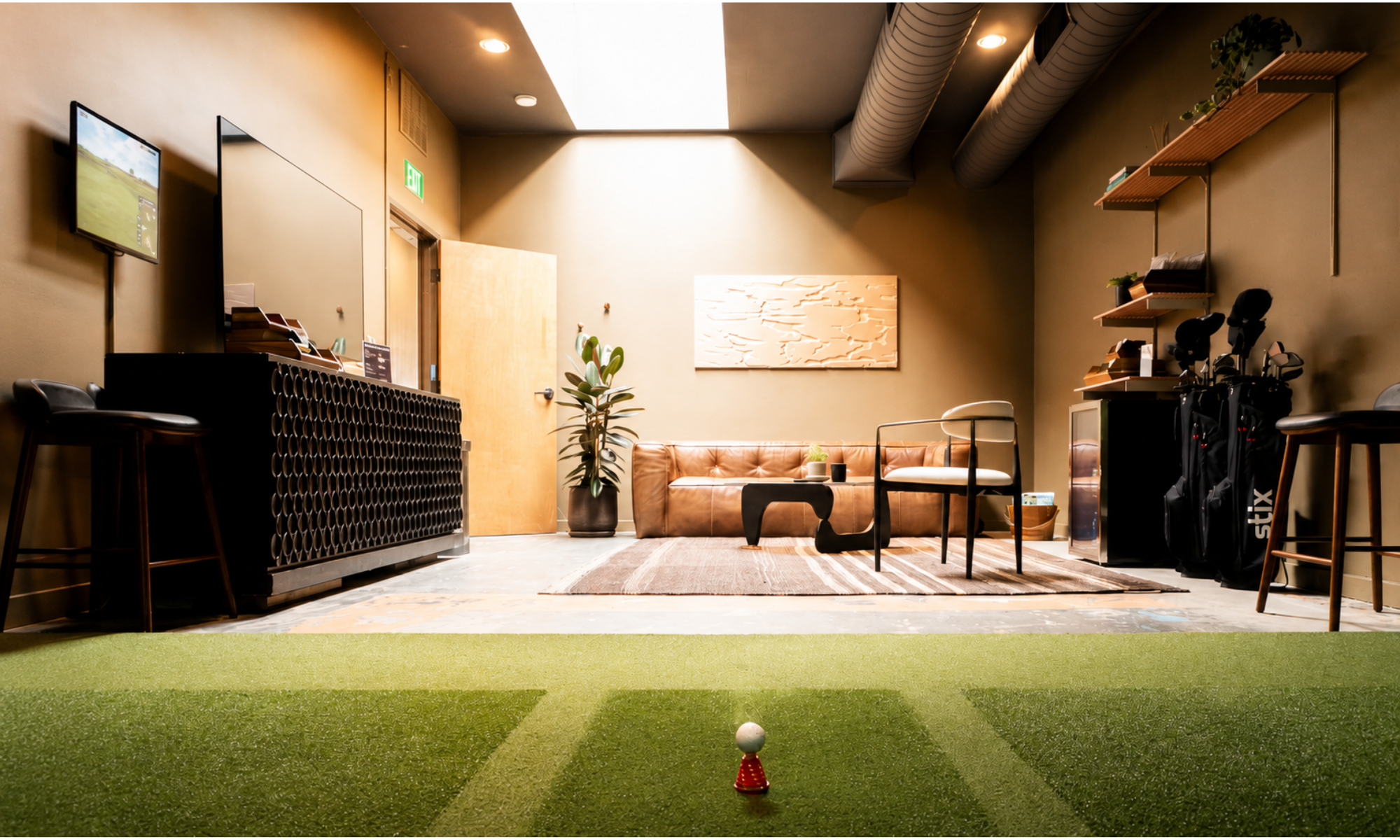 Indoor golf practice room with artificial turf, a golf ball on a tee, seating area with chairs, a leather sofa, a small table, wall decorations, shelves with books and golf equipment, and a golf game displayed on a wall television.