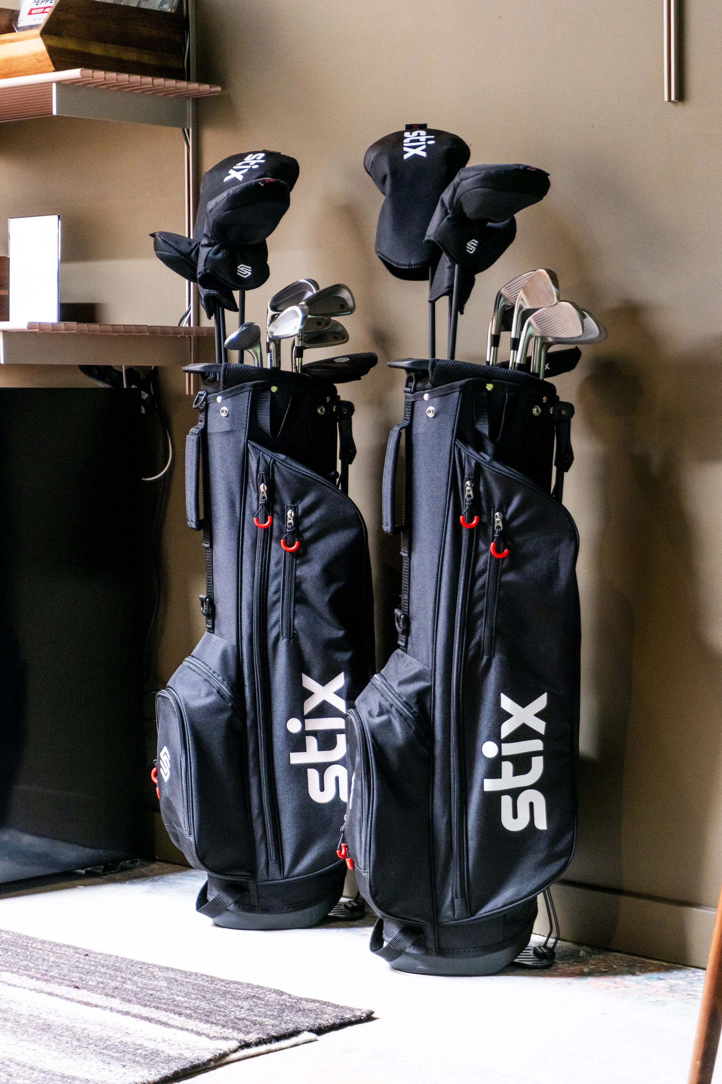 Two black golf bags with golf clubs inside, standing on a beige carpeted floor next to a wall.