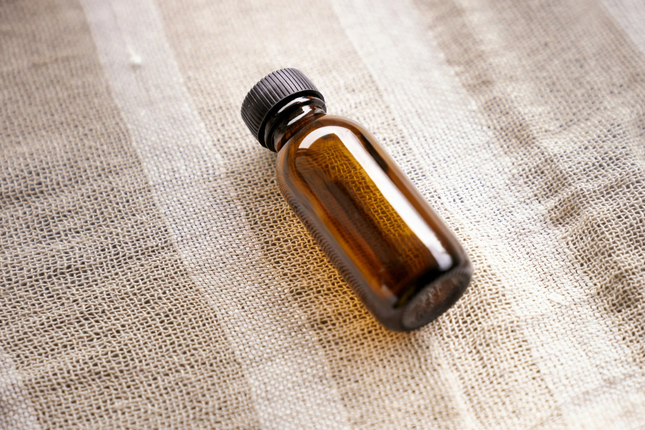 Small amber glass bottle with a black screw cap lying on a textured beige fabric surface.