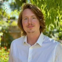 A young man with shoulder-length brown hair and a mustache, wearing a white button-up shirt, outdoors with greenery in the background.