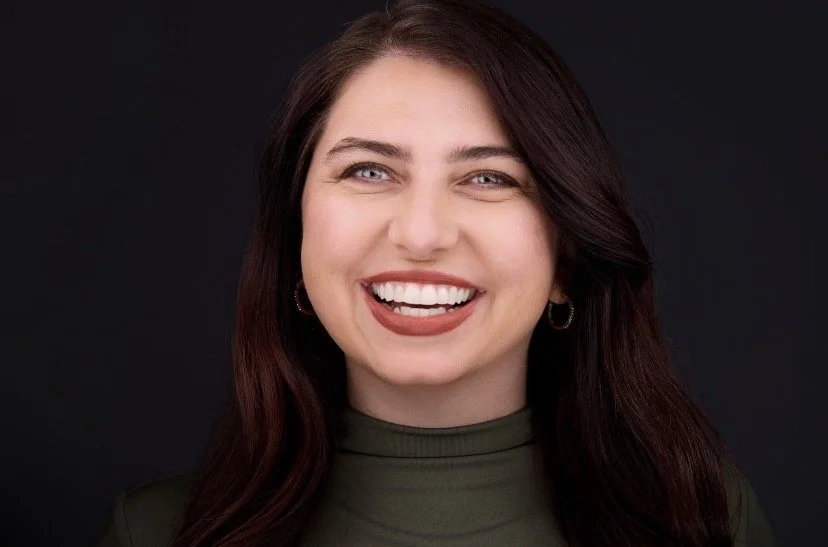 A woman with long dark hair smiling and sticking out her tongue against a black background.