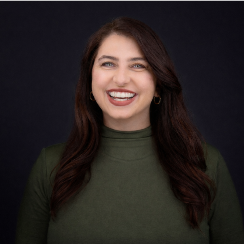 A woman with long, wavy brown hair smiling at the camera, wearing an olive green turtleneck sweater and small hoop earrings, against a dark background.