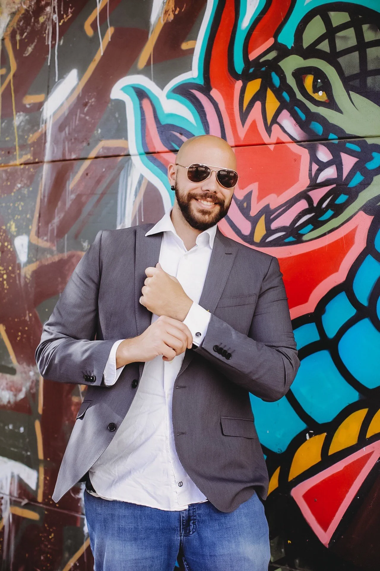 A man with a beard and bald head wearing sunglasses, a gray suit, and a white shirt, standing in front of a colorful graffiti mural on a wall.