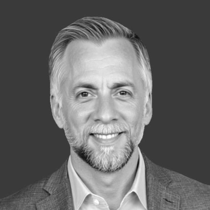 Black and white headshot of a smiling middle-aged man with short, styled hair, a beard, and wearing a suit jacket and collared shirt, against a dark background.