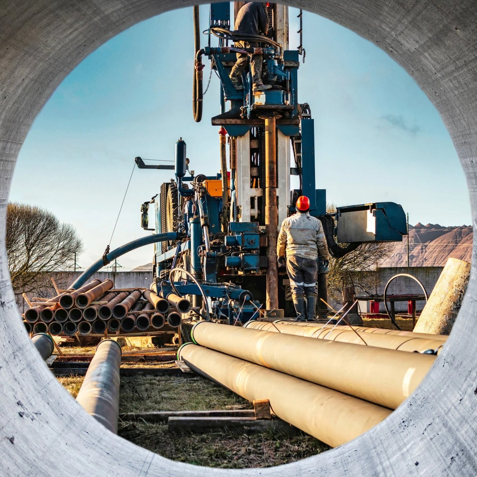 View through a large pipe showing workers operating drilling equipment to install underground pipes or infrastructure.