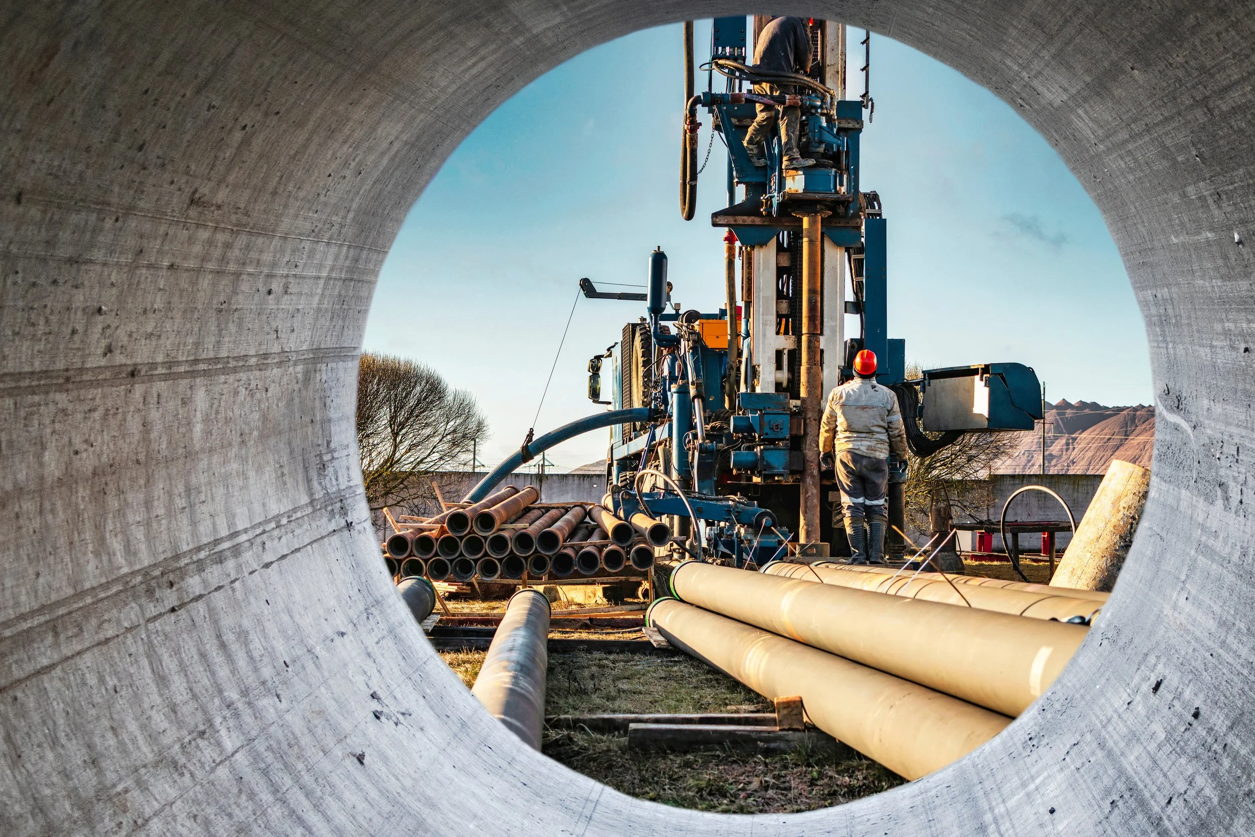 View through a large concrete pipe showing a worker operating heavy machinery and stacked pipes at an industrial site during daytime.