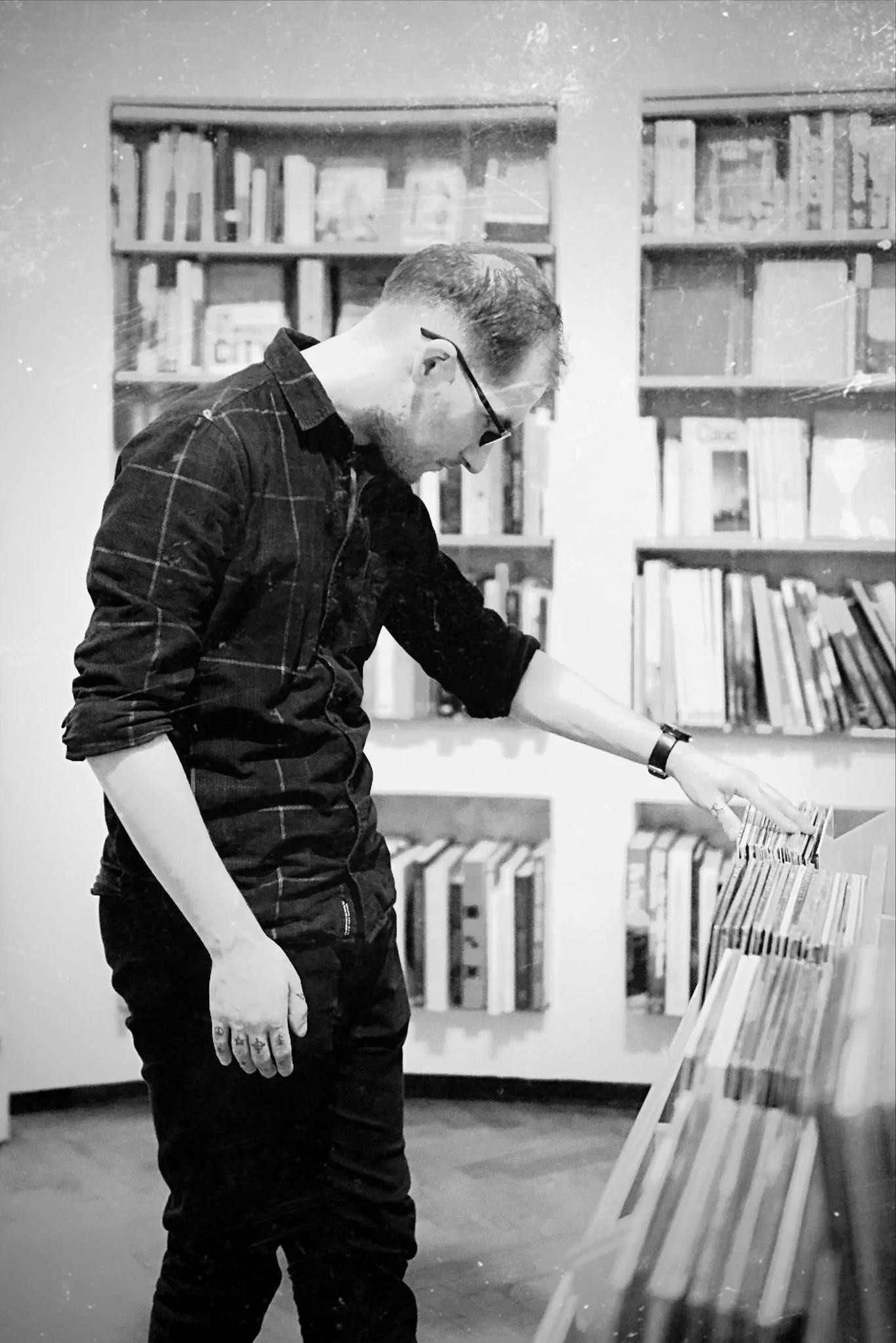 A man wearing glasses and a patterned shirt is browsing books in a bookstore or library, with shelves of books in the background.