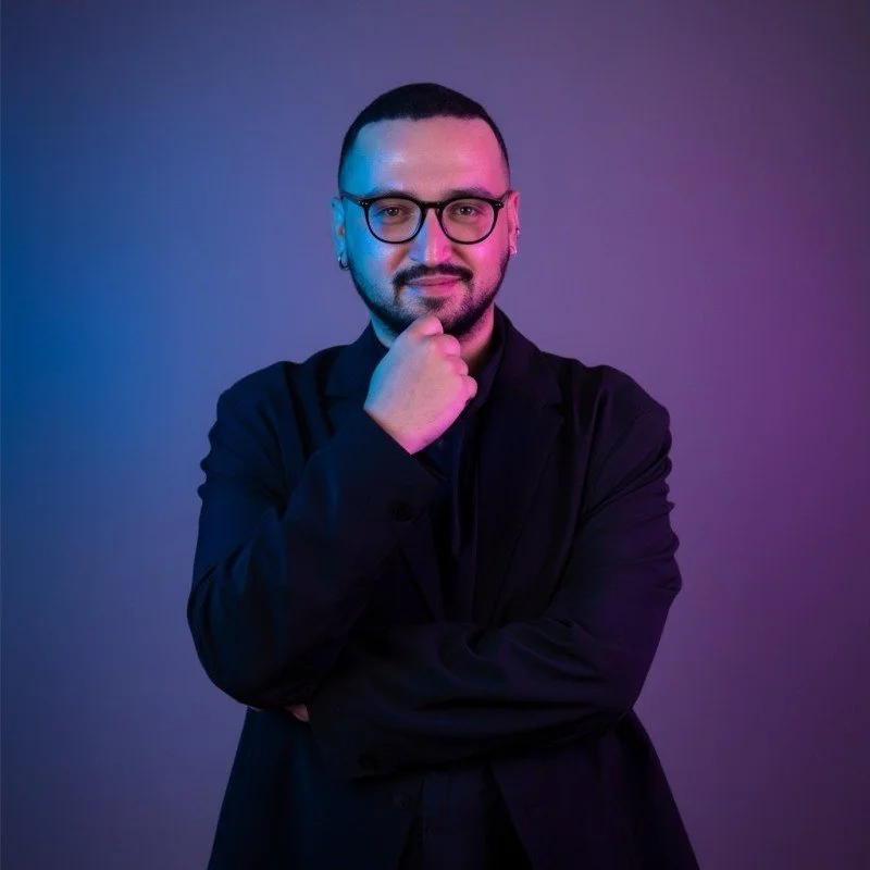 A man with glasses, a beard, and short hair posing confidently with his hand on his chin against a purple and blue gradient background.
