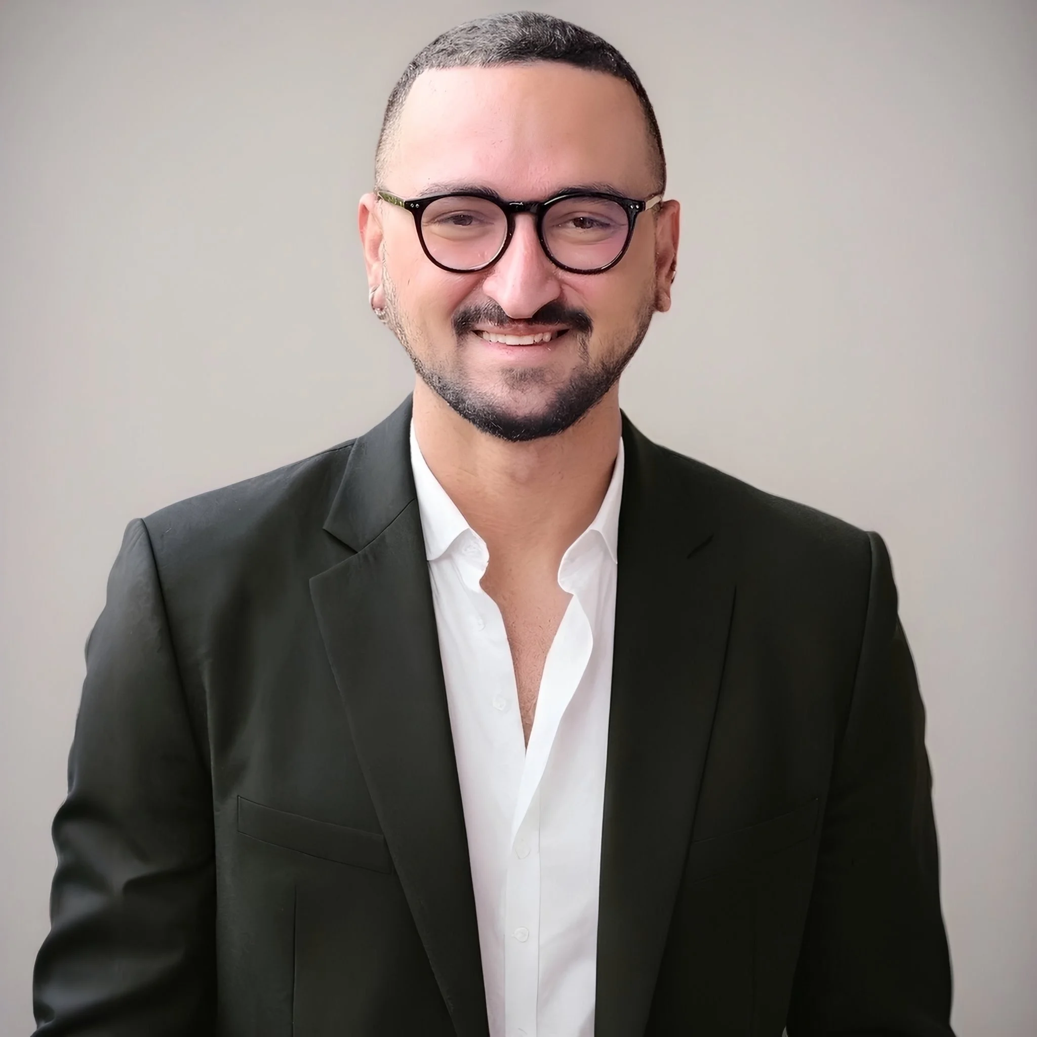 A man with short hair, glasses, and a beard, wearing a black suit and white shirt, smiling and looking at the camera against a plain light-colored background.