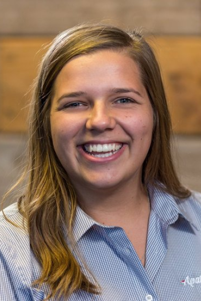 Close-up of a smiling young woman with light brown hair wearing a blue collared shirt in front of a wooden background.