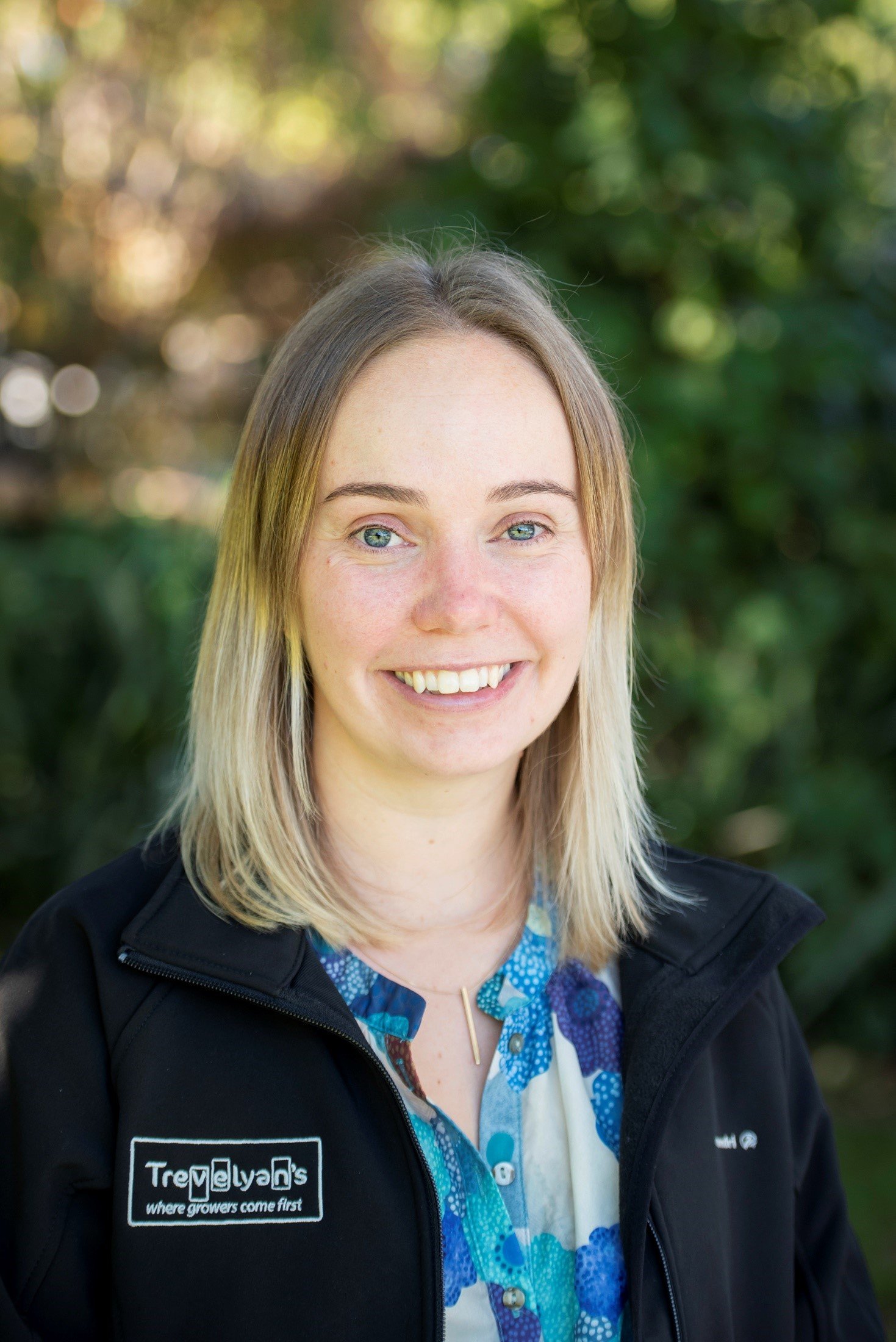 A woman with blonde hair and blue eyes smiling outdoors, wearing a black jacket with a patch that reads 'Trevelyan's where growers come first' and a colorful blue patterned shirt.