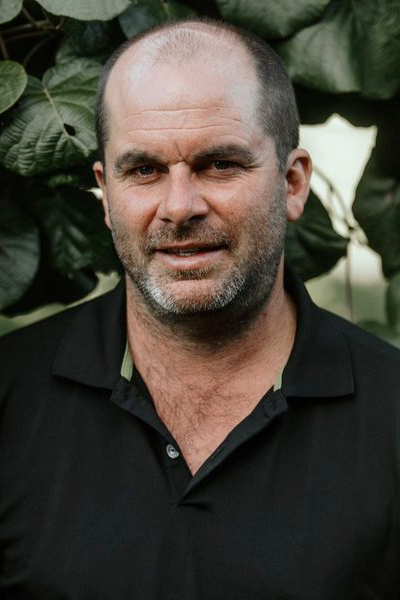 A middle-aged man with a shaved head and beard, wearing a black collared shirt, standing outdoors in front of large green leaves.