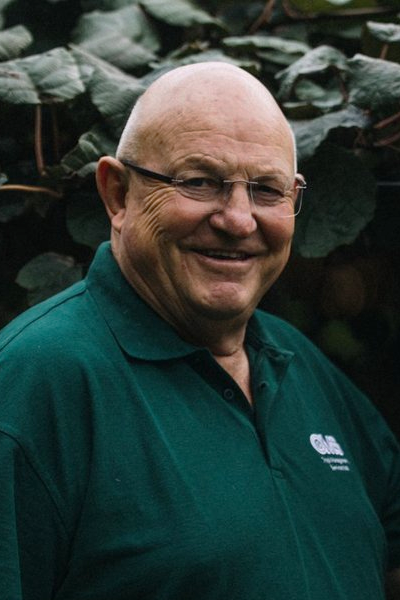 A smiling, bald man wearing glasses and a teal polo shirt standing outdoors in front of large green leaves.