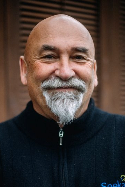 A smiling middle-aged man with a bald head and a gray beard, wearing a black zip-up sweater, standing in front of wooden shutters.